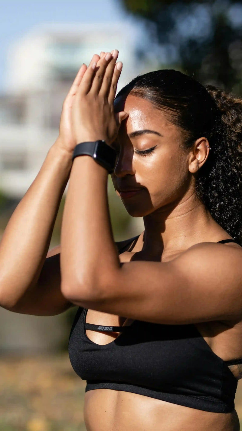 Woman in black sports bra and shorts standing outdoors with hands on head, blurred background